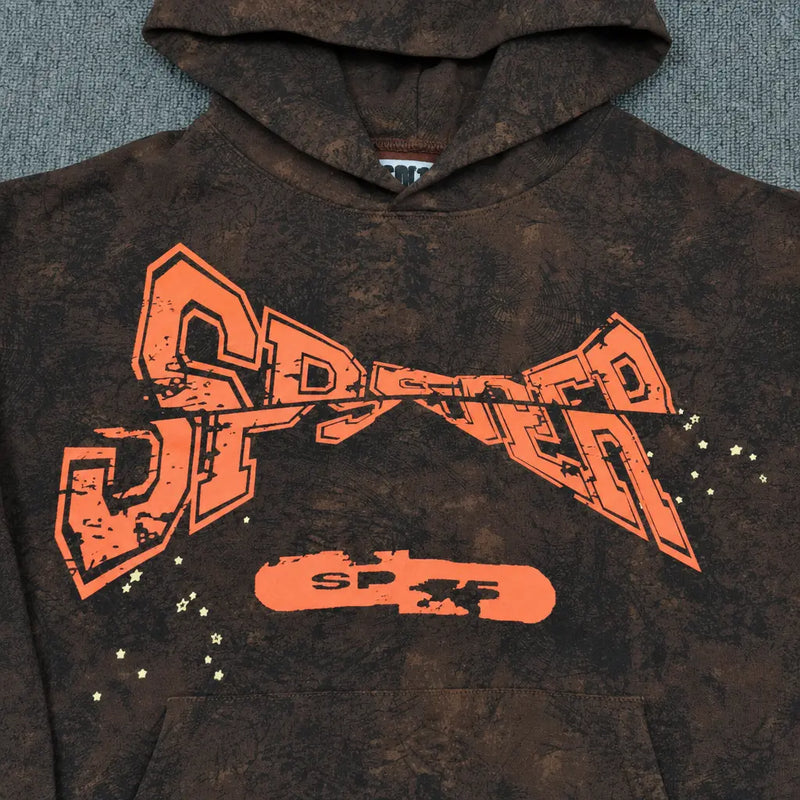 The Sp5der Brown Tie-Dye Hoodie with Orange Logo Print in streetwear style features bold orange SPSTER text, star accents, and a skateboard graphic, displayed on a textured gray surface.