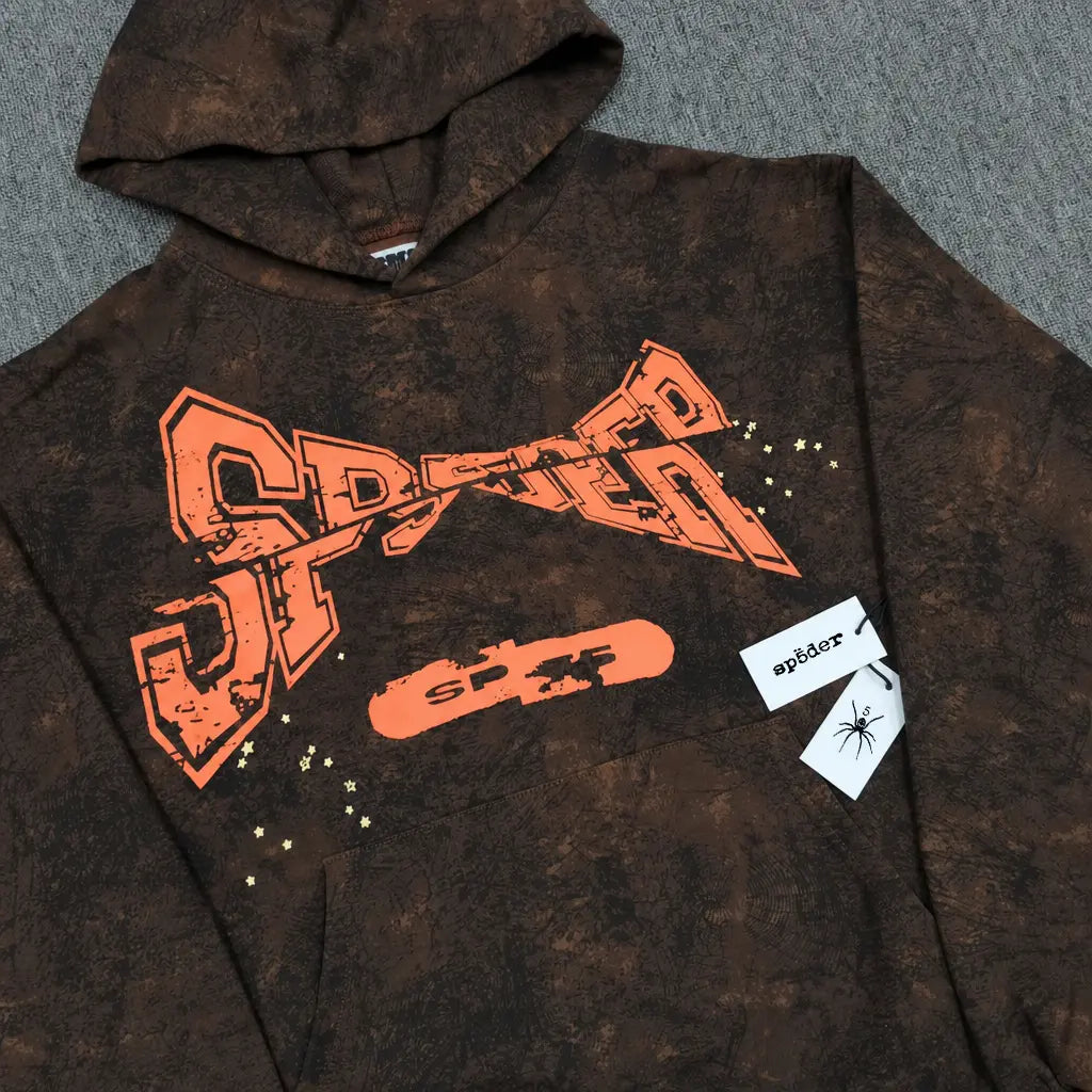 The Sp5der Brown Tie-Dye Hoodie with Orange Logo Print features a bold orange SP5DER logo, skateboard graphic, star accents, and mottled texture. Two tags show "sp5der" and a spider illustration—perfect for standout streetwear style.