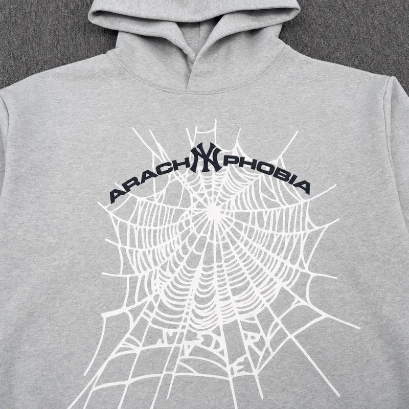 The Sp5der Arachnophobia NY Spiderweb Graphic Hoodie Grey features a striking white spiderweb and curved black ARACHNOPHOBIA text on the front, laid flat on a dark textured surface for a modern streetwear appeal.
