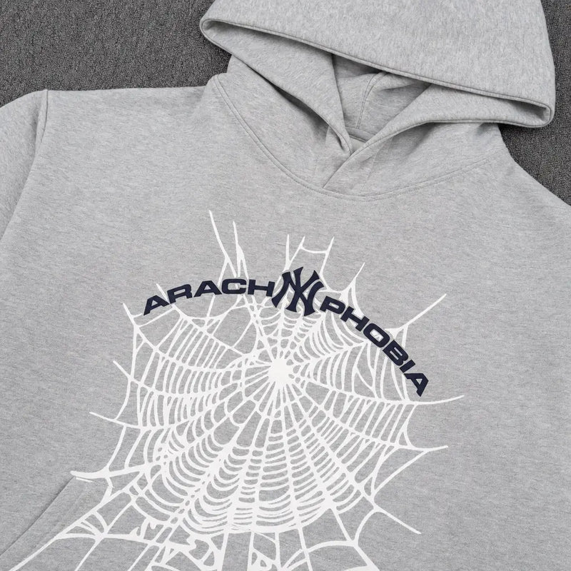 The Sp5der Arachnophobia NY Spiderweb Graphic Hoodie Grey features a bold spiderweb design, dark blue ARACHNOPHOBIA text above, and a stylized NY logo at the center—a standout piece for any streetwear collection.