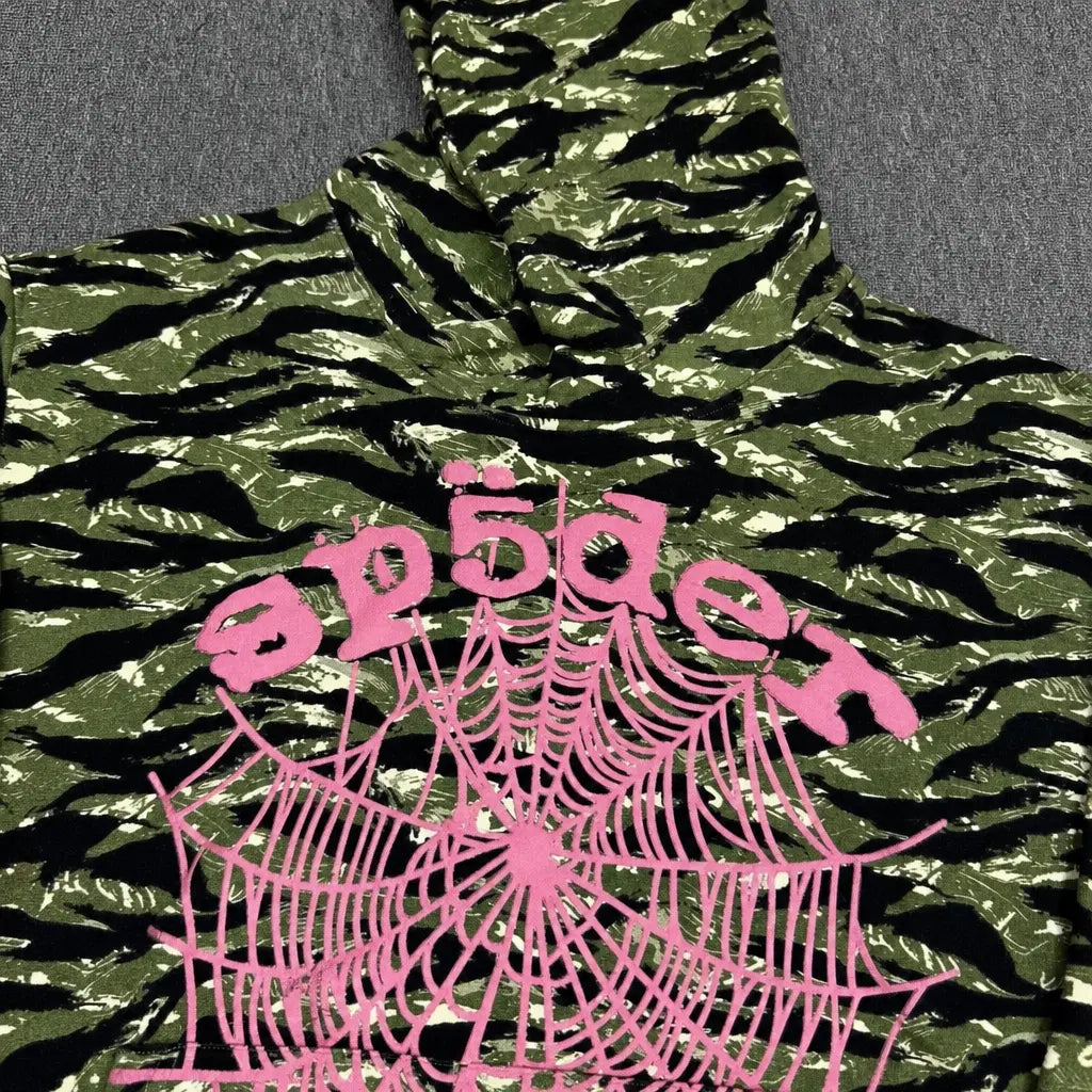 The Sp5der Tiger Camo Hoodie with Pink Spiderweb Logo showcases a green tiger camo and black design, bold pink spiderweb graphic, and distorted pink "sp5der" text—making it a standout piece for any streetwear collection.
