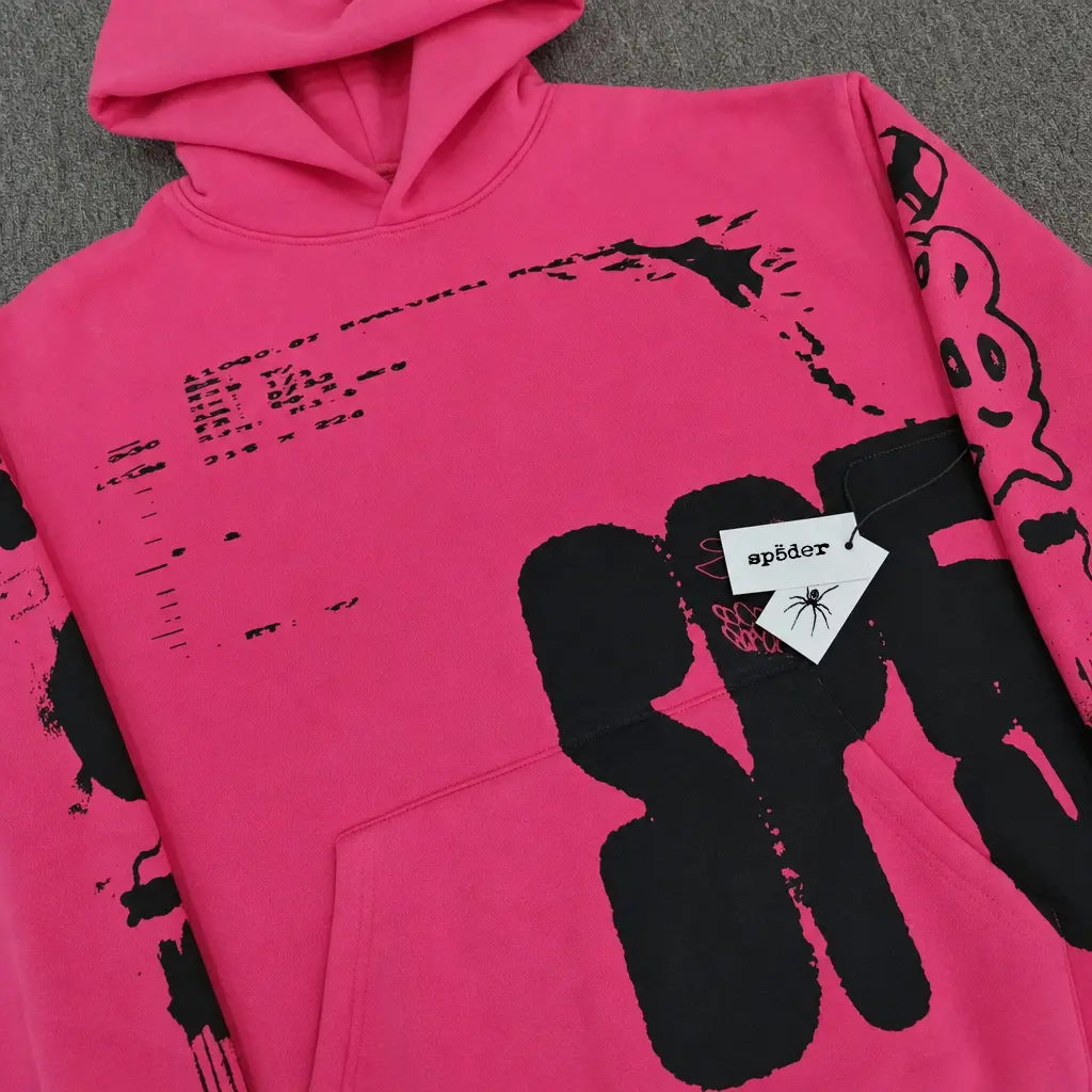 The Sp5der Hot Pink Hoodie Black Graffiti Text Print features bold graphics and a signature "sp5der" tag on the front pocket, laid flat on a grey surface—an ideal choice for any streetwear collection.