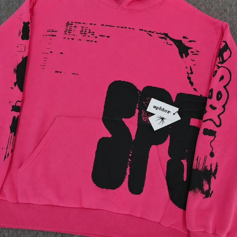 The Sp5der Hot Pink Hoodie Black Graffiti Text Print features abstract black lettering, a white sp5der tag with a spider graphic on the front pocket, and is displayed flat on a gray surface.