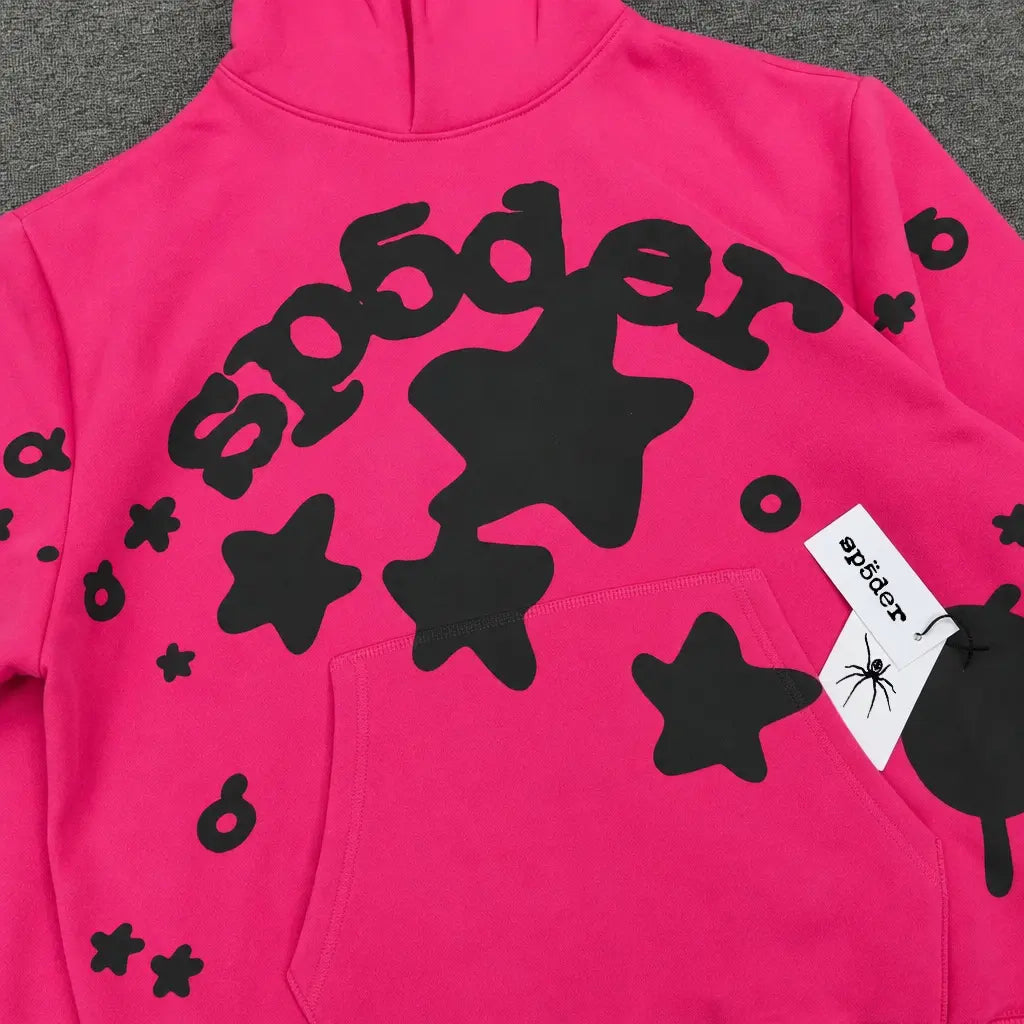 The Sp5der Hot Pink Black Star Logo Print Hoodie features bold black stars, "sp5der" stylized across the chest, and a white tag with sp5der and spider graphic on the pocket—ideal for streetwear fans.