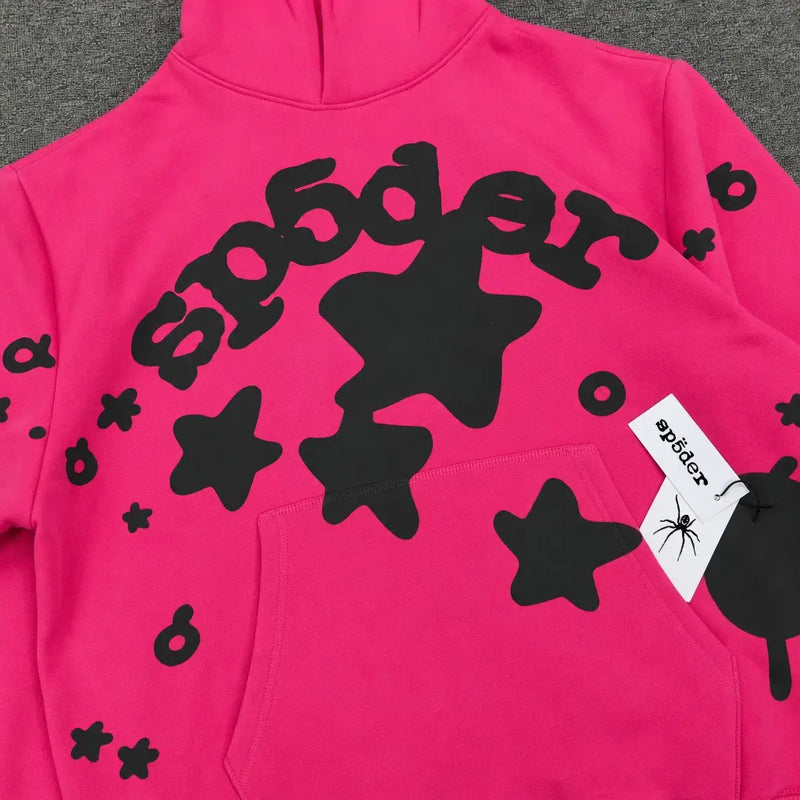 The Sp5der Hot Pink Black Star Logo Print Hoodie features bold black stars, "sp5der" stylized across the chest, and a white tag with sp5der and spider graphic on the pocket—ideal for streetwear fans.