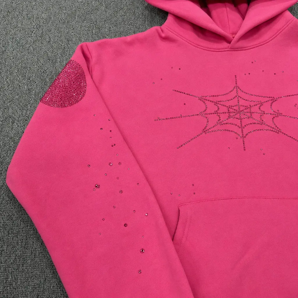 The Sp5der Pink Rhinestone Spiderweb Crystal Hoodie features a hot pink design with rhinestone spiderwebs on the chest, sleeve accents, and a glittery elbow patch—making it the ultimate statement streetwear pullover.