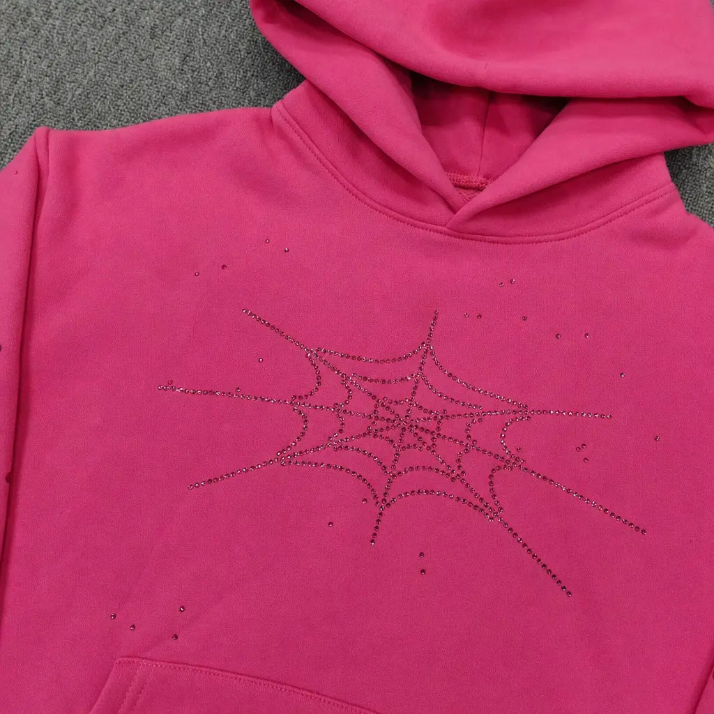 The Sp5der Pink Rhinestone Spiderweb Crystal Hoodie features a sparkling spiderweb design on hot pink, displayed on a gray textured surface—an eye-catching addition to any streetwear pullover collection.