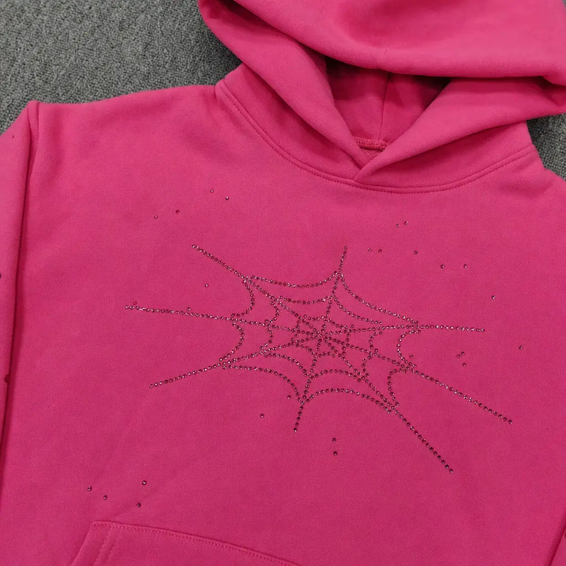 The Sp5der Pink Rhinestone Spiderweb Crystal Hoodie features a sparkling spiderweb design on hot pink, displayed on a gray textured surface—an eye-catching addition to any streetwear pullover collection.