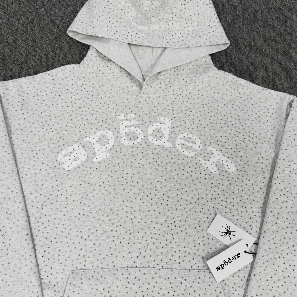 The Sp5der Light Grey Rhinestone All-Over Arch Logo Hoodie features a textured dotted pattern and a stylized white "sp5der" print across the chest, with two branded tags attached for authentic streetwear appeal.