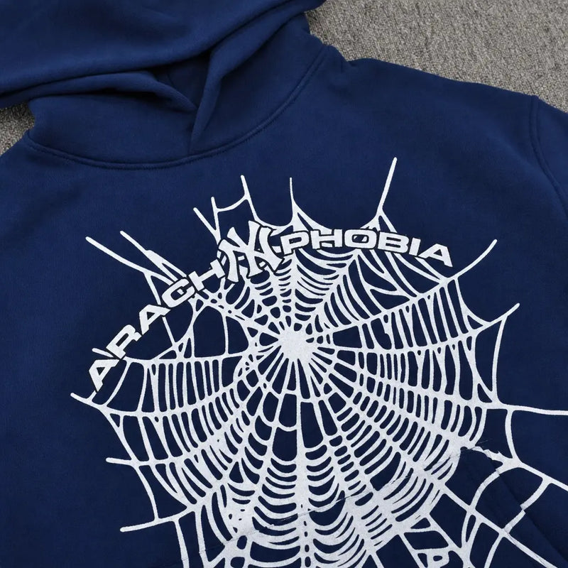The Sp5der Arachnophobia Navy Blue Spiderweb Print Hoodie features a bold white spiderweb and "ARACHNOPHOBIA" across the back, shown laid flat on a textured gray surface.