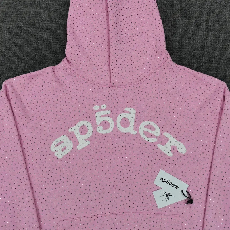 The Sp5der Pink Rhinestone All-Over Arch Logo Hoodie features small black dots, bold white “sp5der” text across the chest, and a tag with the sp5der logo and a spider graphic, making it a stylish streetwear piece.