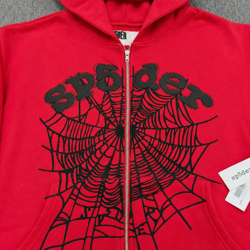 The Sp5der Red Zip-Up Hoodie Black Spiderweb Studded Logo features a bold black spiderweb and "sp5der" lettering on the front, plus a signature white tag on the zipper—making it a standout streetwear essential.