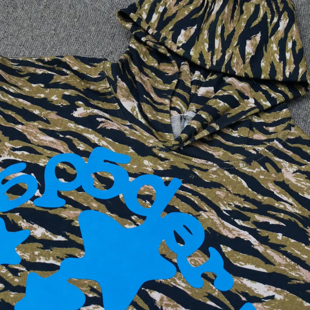 A close-up of the Sp5der Tiger Camo Blue Star Print Logo Hoodie, featuring a green, brown, and black camo design with bold blue lettering and shapes on the front, laid flat on a gray surface.