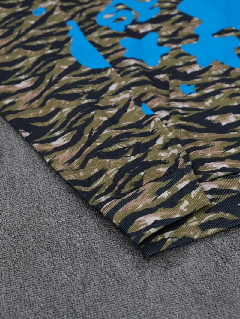 Close-up of the Sp5der Tiger Camo Blue Star Print Logo Hoodie, showcasing its green and brown tiger camo with blue star accents, laid on a gray textured surface.