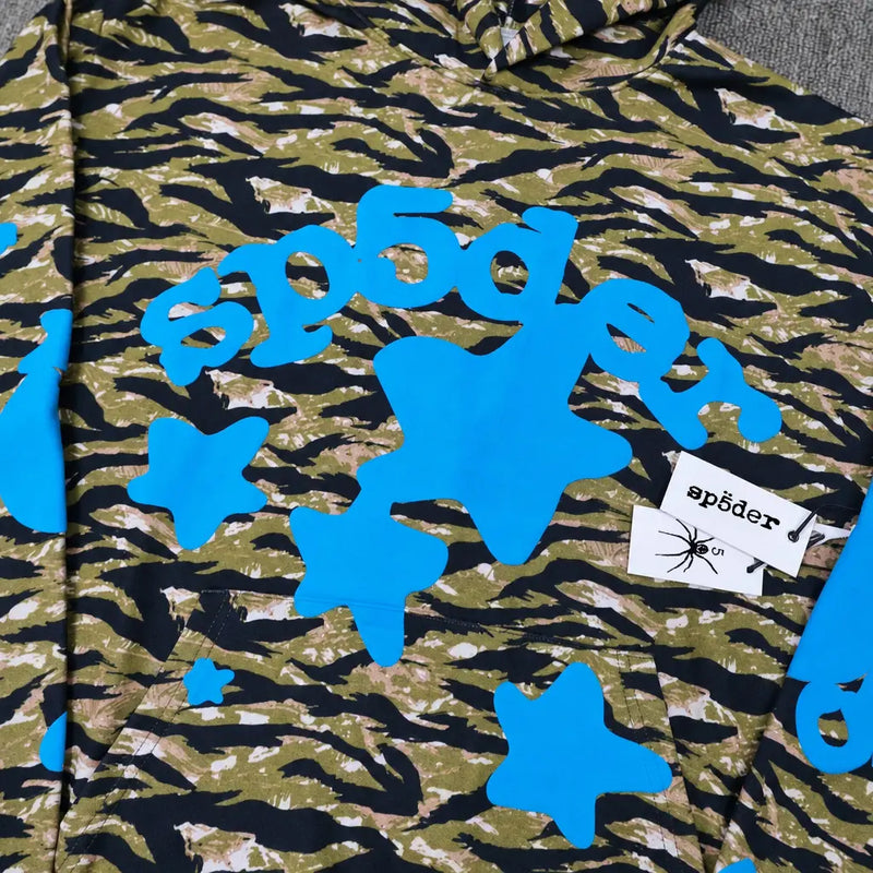 The Sp5der Tiger Camo Blue Star Print Logo Hoodie features a green and black tiger camo pattern, blue stars, bold blue “sp5der” text, and two white tags—one with the sp5der logo and a spider graphic.