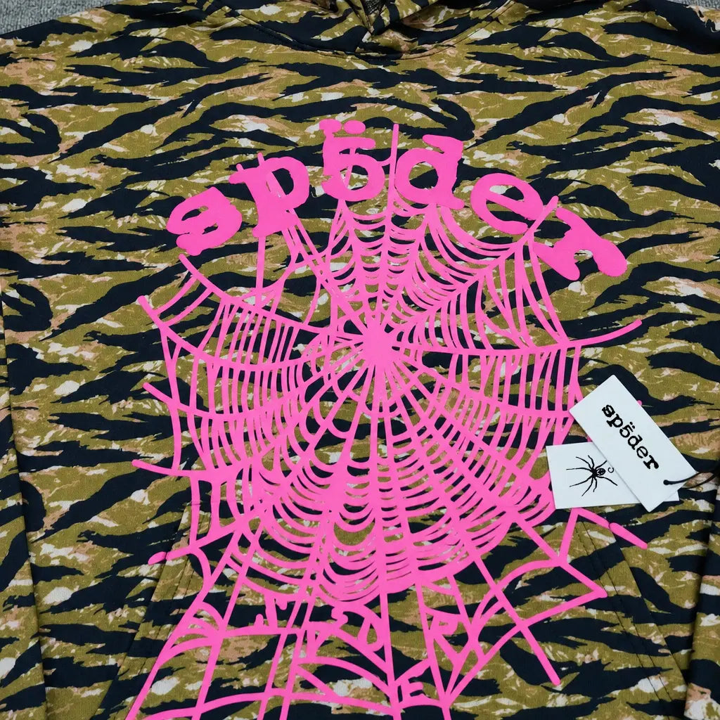 The Sp5der Tiger Camo Hoodie features a streetwear-inspired camouflage pattern, a large pink spiderweb print with "sp5der" in pink text, and two attached white tags—one displaying the sp5der logo and a black spider graphic.