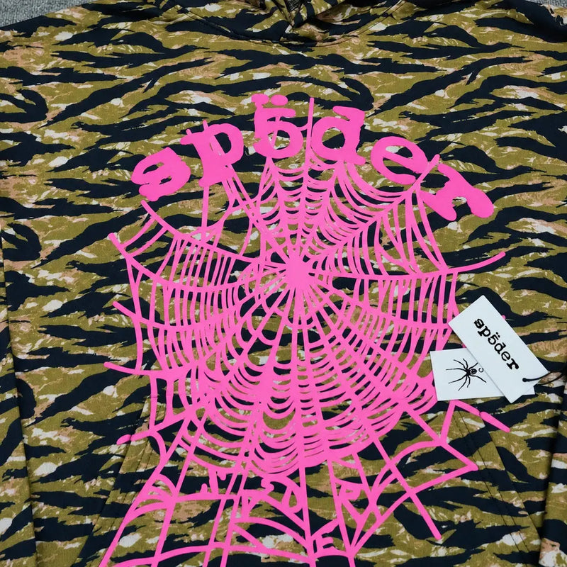 The Sp5der Tiger Camo Hoodie features a streetwear-inspired camouflage pattern, a large pink spiderweb print with "sp5der" in pink text, and two attached white tags—one displaying the sp5der logo and a black spider graphic.