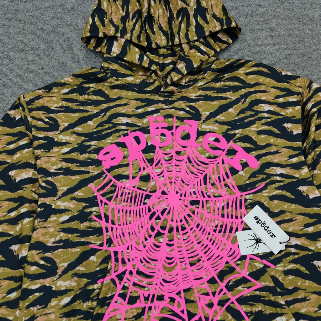 The Sp5der Tiger Camo Hoodie with Pink Spiderweb Print showcases bold brown and black stripes, a vivid pink spiderweb graphic, "spider" on the front, and a white tag featuring Sp5der branding with a spider illustration.