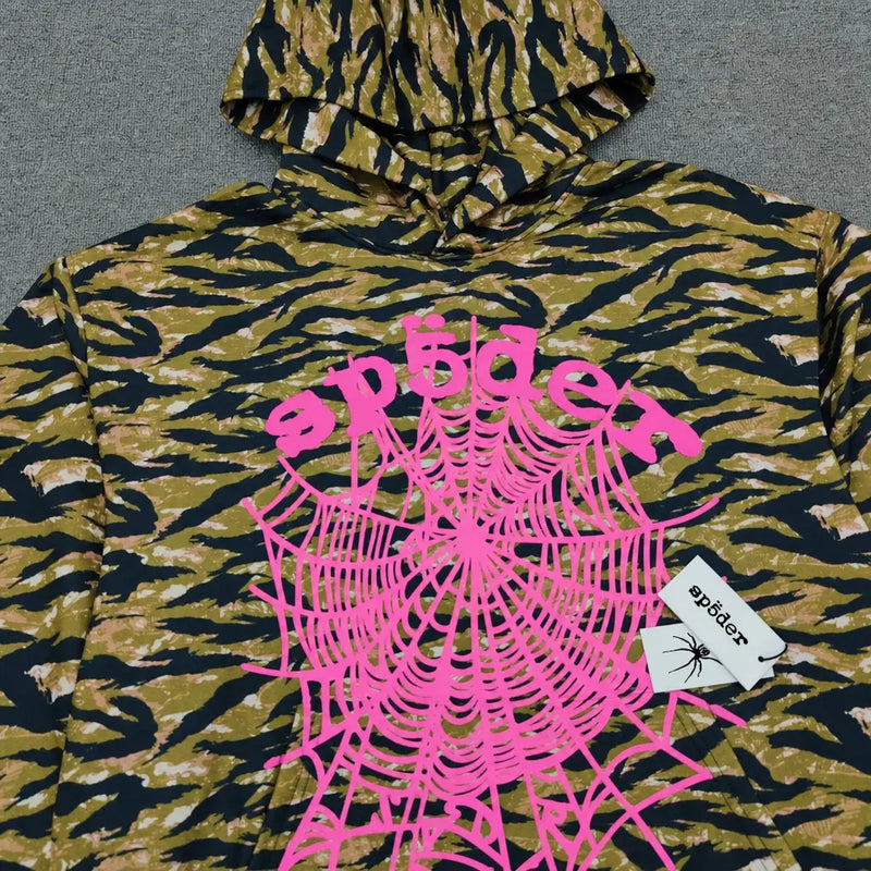 The Sp5der Tiger Camo Hoodie with Pink Spiderweb Print showcases bold brown and black stripes, a vivid pink spiderweb graphic, "spider" on the front, and a white tag featuring Sp5der branding with a spider illustration.