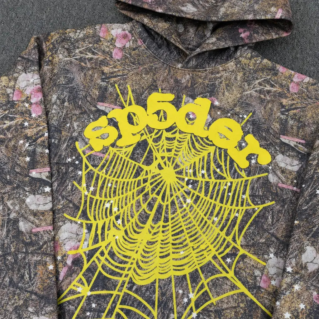 The Sp5der Woodland Camo Floral Web Graphic Hoodie features a camo pattern, bold yellow “spōder” lettering, and a striking spiderweb graphic—delivering standout streetwear style to your collection.