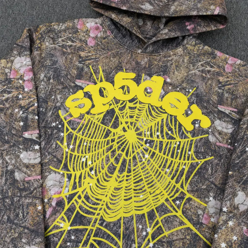 The Sp5der Woodland Camo Floral Web Graphic Hoodie features a camo pattern, bold yellow “spōder” lettering, and a striking spiderweb graphic—delivering standout streetwear style to your collection.