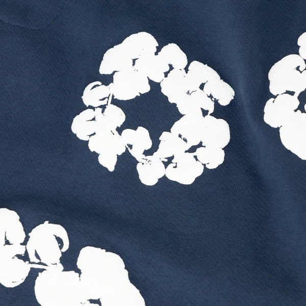 The Navy Blue “Cotton Wreath” Denim Tears Sweatpants feature white, irregular floral wreath prints in circular patterns, offering a unique streetwear style.