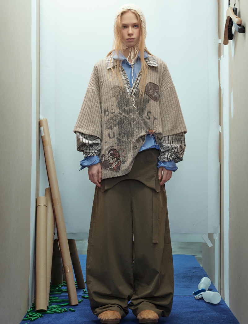 Model wearing beige layered bear graphic sweater with plaid sleeves and wide olive pants