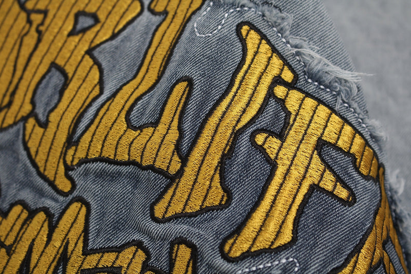 Close-up of gray vintage washed denim fabric with frayed edges and gold embroidered graphic stitching on a distressed T-shirt