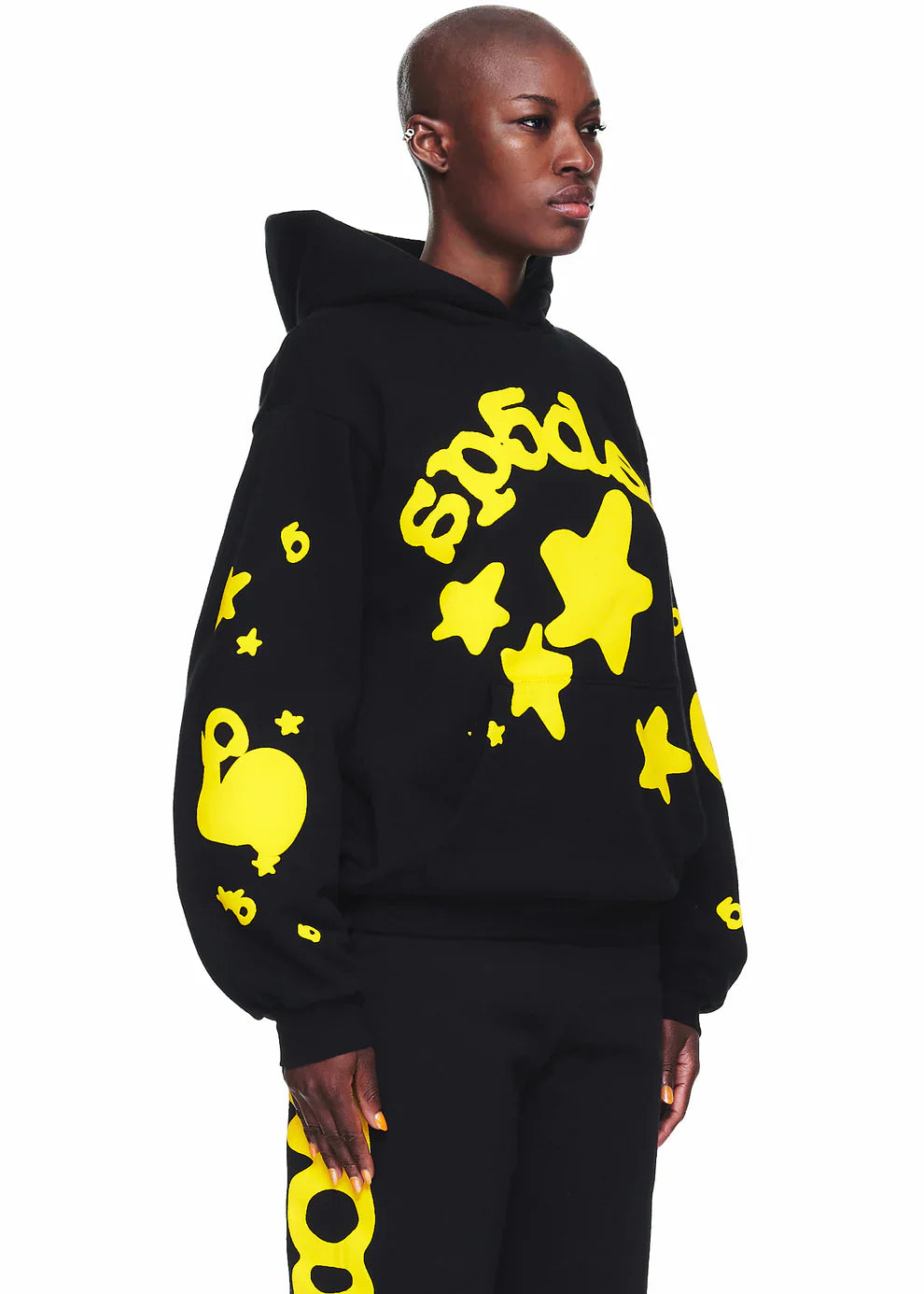 A person in the Black Graphic Hoodie with Star Lettering Print stands facing left against a white background, pairing the comfort fit pullover with matching pants featuring bold yellow star and abstract designs for a modern streetwear look.