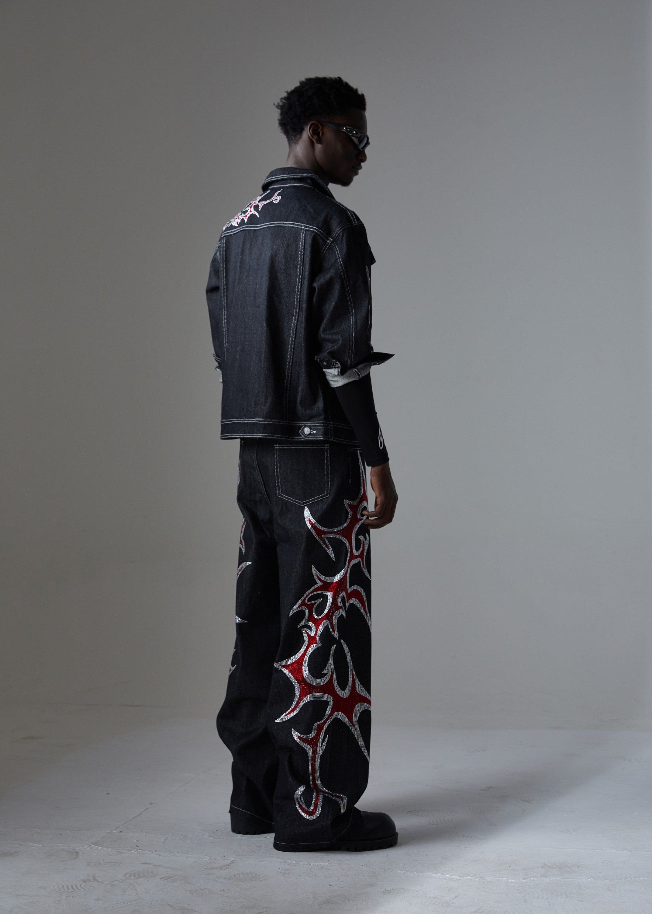 Man wearing black graphic denim jacket with red and white tribal embroidery, styled with matching wide-leg denim trousers