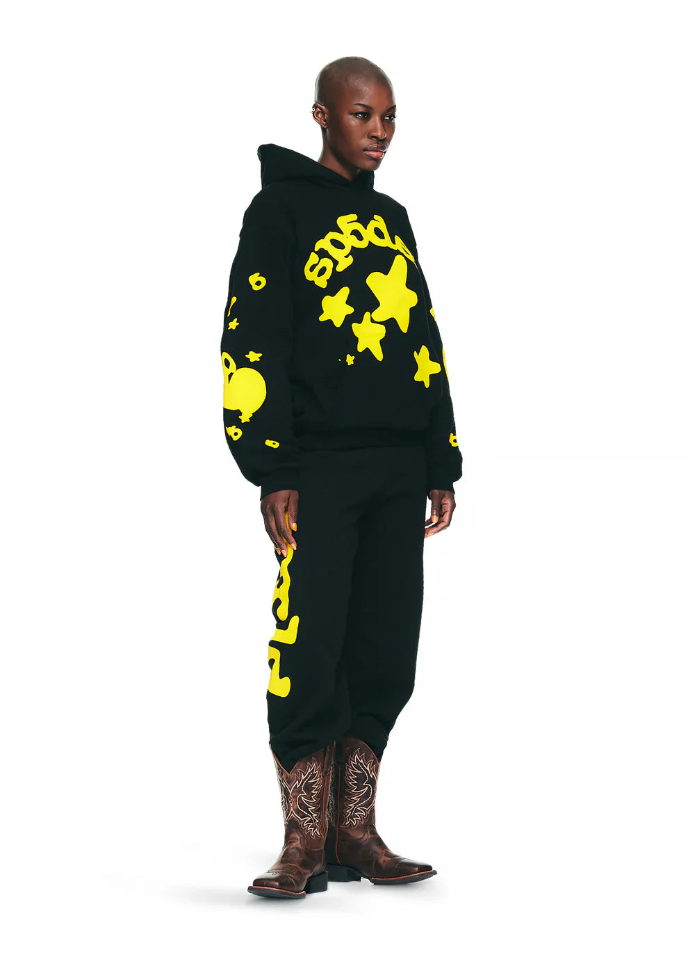 A person stands facing forward in a Black Graphic Hoodie with Star Lettering Print – Comfort Fit Pullover Hoodie for Everyday Wear, paired with sweatpants featuring yellow star designs and brown embroidered cowboy boots, against a white background.