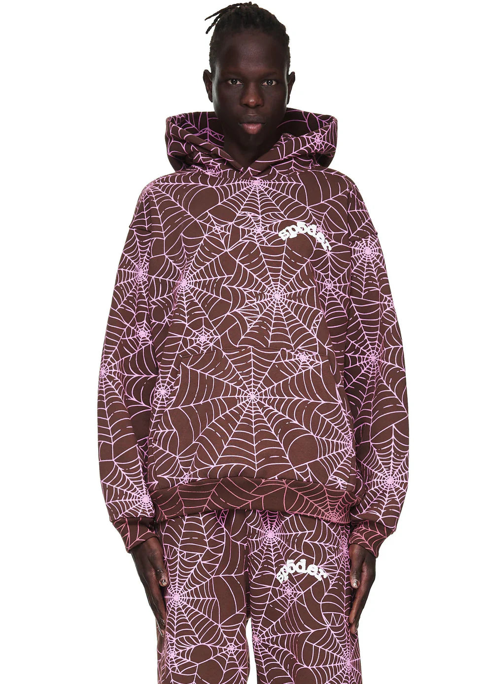 Wearing the Brown All-Over Spider Web Graphic Hoodie – Comfort Fit Pullover, featuring bold pink web prints and a white logo on the chest, a person stands facing forward with the hood up, paired with matching graphic pants.