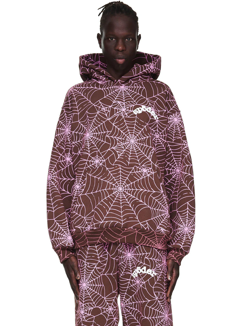 Wearing the Brown All-Over Spider Web Graphic Hoodie – Comfort Fit Pullover, featuring bold pink web prints and a white logo on the chest, a person stands facing forward with the hood up, paired with matching graphic pants.