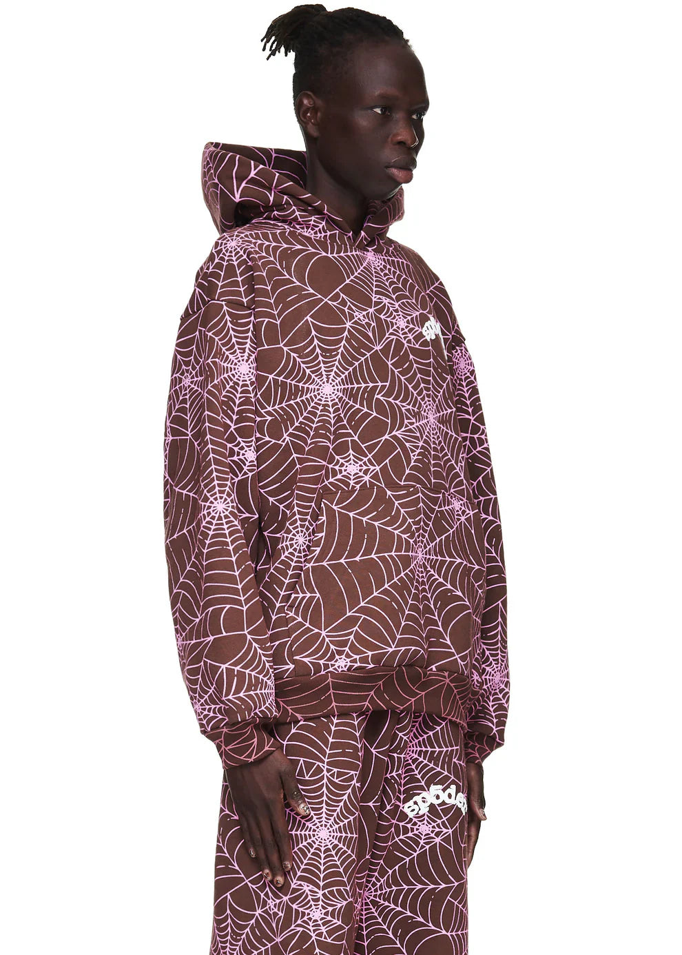 A person wears the Brown All-Over Spider Web Graphic Hoodie – Comfort Fit Pullover Hoodie in brown with a pink spiderweb pattern, styled with matching pants, standing sideways and looking forward.