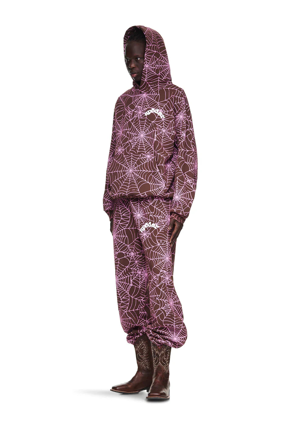 A person wears the Brown All-Over Spider Web Graphic Hoodie – Comfort Fit Pullover with matching sweatpants, both featuring a pink spiderweb pattern and white logo, paired with brown cowboy boots against a white background.