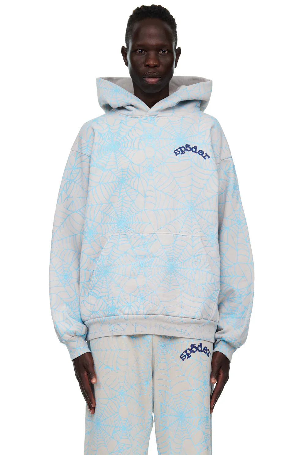 A person wears the Light Grey All-Over Spider Web Graphic Hoodie – Comfort Fit Pullover Hoodie and matching pants, both featuring bold blue spider web patterns and “Sp5der” branding, standing against a white background.
