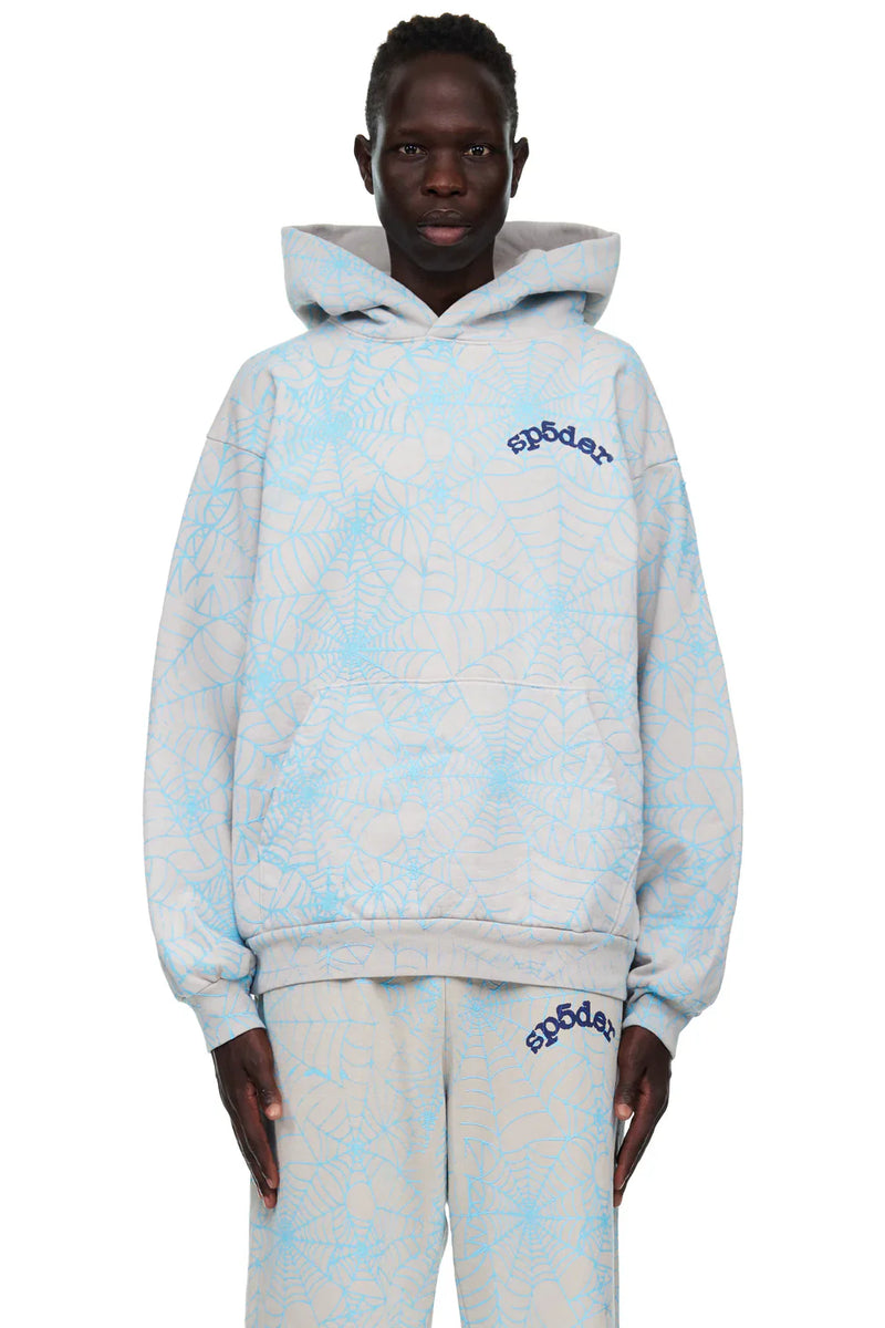 A person wears the Light Grey All-Over Spider Web Graphic Hoodie – Comfort Fit Pullover Hoodie and matching pants, both featuring bold blue spider web patterns and “Sp5der” branding, standing against a white background.