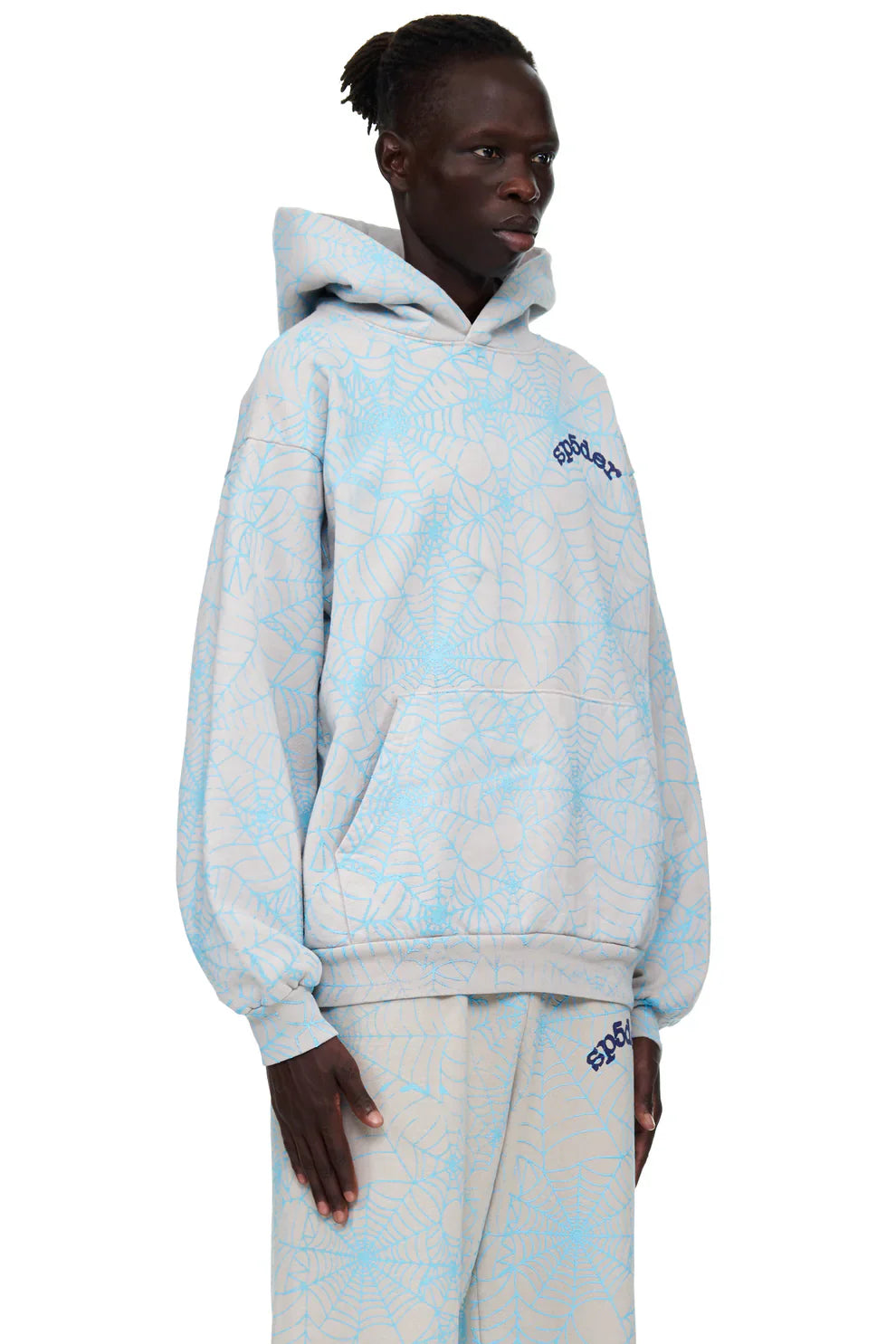 A person stands against a white background wearing the Light Grey All-Over Spider Web Graphic Hoodie—Comfort Fit Pullover Hoodie—and matching pants, both featuring blue web-like patterns and blue text on the chest and thigh.