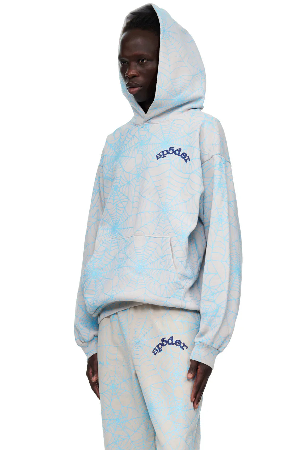 A person wearing the Light Grey All-Over Spider Web Graphic Hoodie – Comfort Fit Pullover Hoodie with matching sweatpants, featuring spider web prints and "sp5der" branding, stands with hands by their sides.