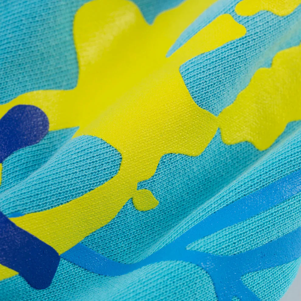 Close-up of the Turquoise Graphic Hoodie with Star Detail and Spider Web Back Print, showing its textured blue fabric with abstract yellow and dark blue shapes, soft folds, and a slightly raised surface; comfort fit pullover.