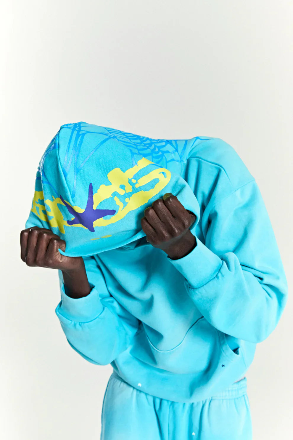 A person in a bright blue tracksuit pulls the hood of their Turquoise Graphic Hoodie with Star Detail and Spider Web Back Print over their head, partially covering their face. The comfort fit pullover features yellow and purple graphic designs on top.