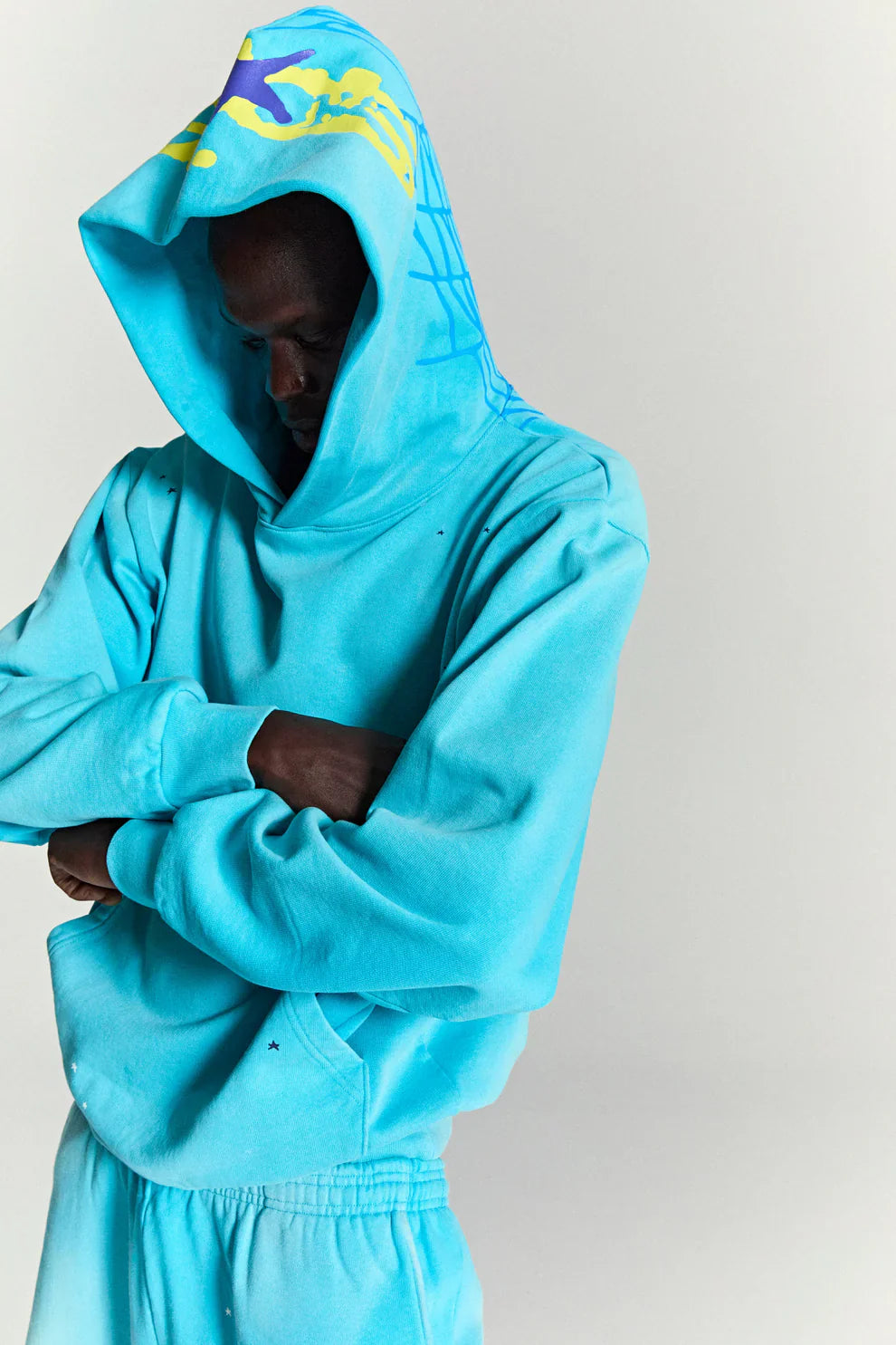 A person in a Turquoise Graphic Hoodie with Star Detail and Spider Web Back Print stands with arms crossed and head bowed against a plain, light background.