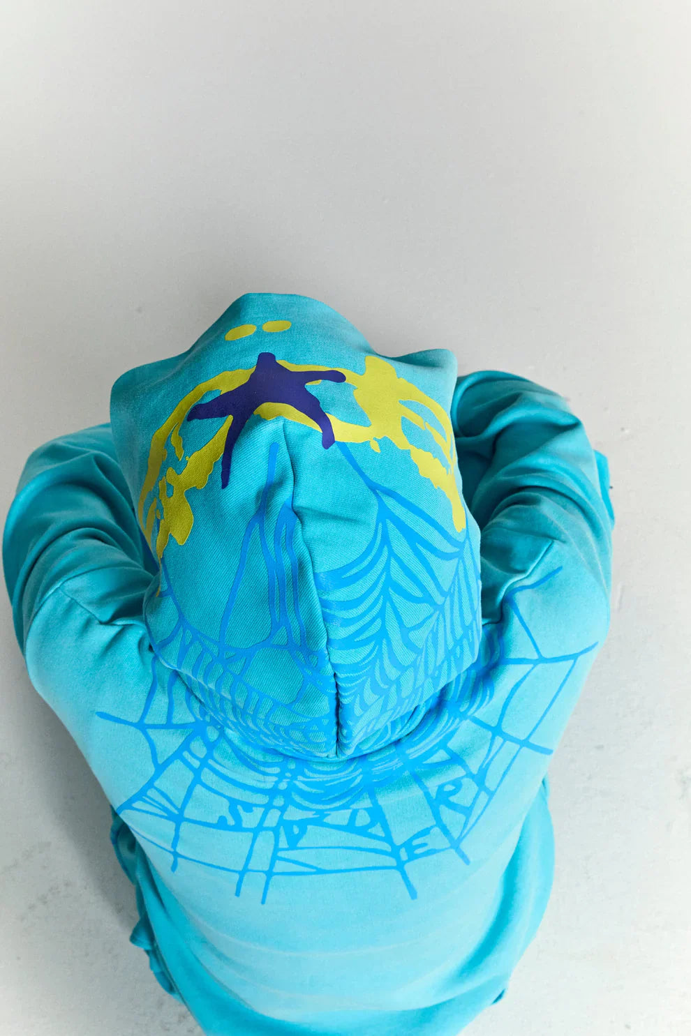 A person wearing the Turquoise Graphic Hoodie with Star Detail and Spider Web Back Print, hood up, is viewed from above while sitting against a light background.