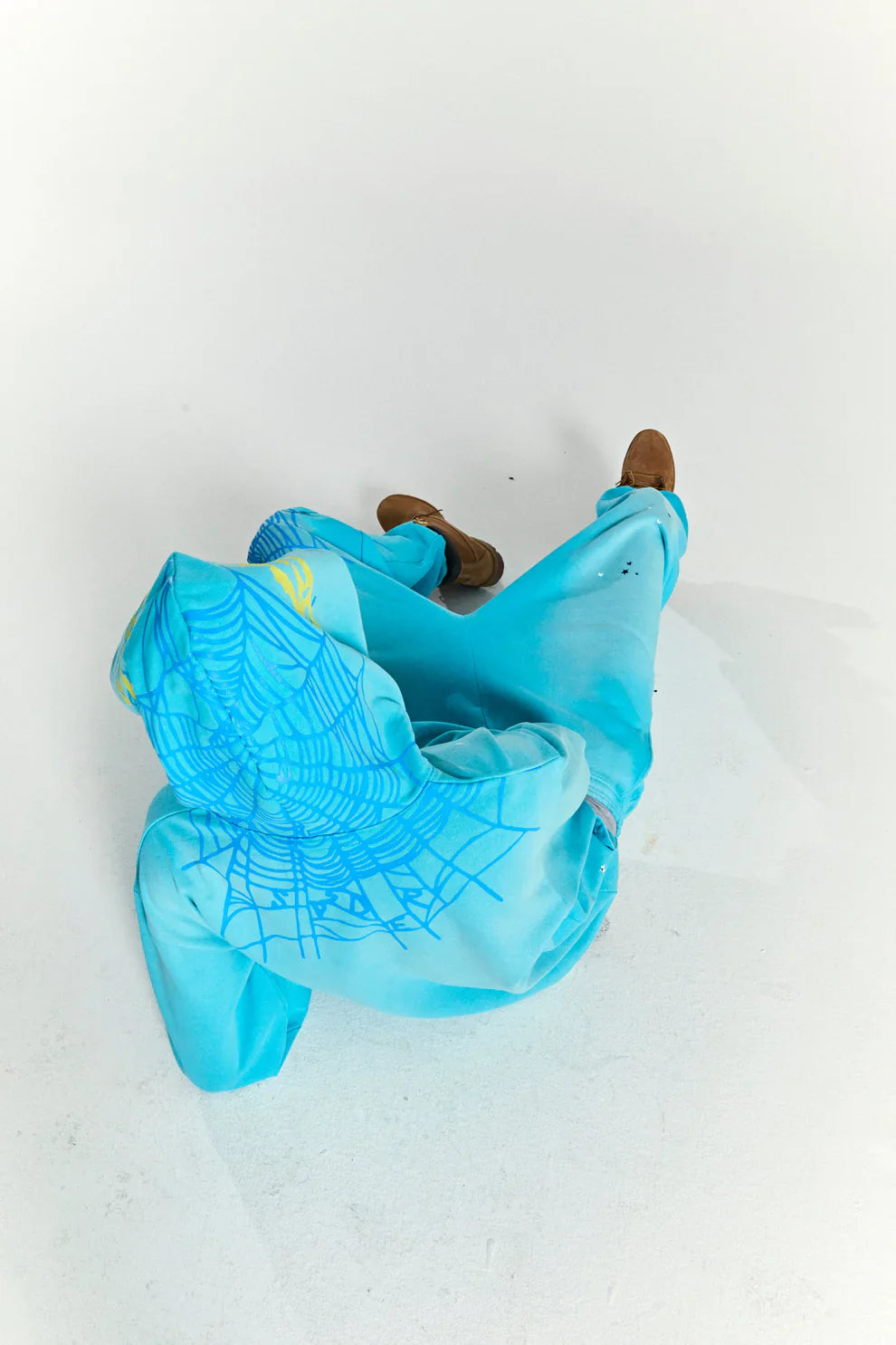 A person wearing a Turquoise Graphic Hoodie with Star Detail and Spider Web Back Print – Comfort Fit Pullover Hoodie sits on a white floor, legs extended, hood up hiding their face, with brown shoes visible.