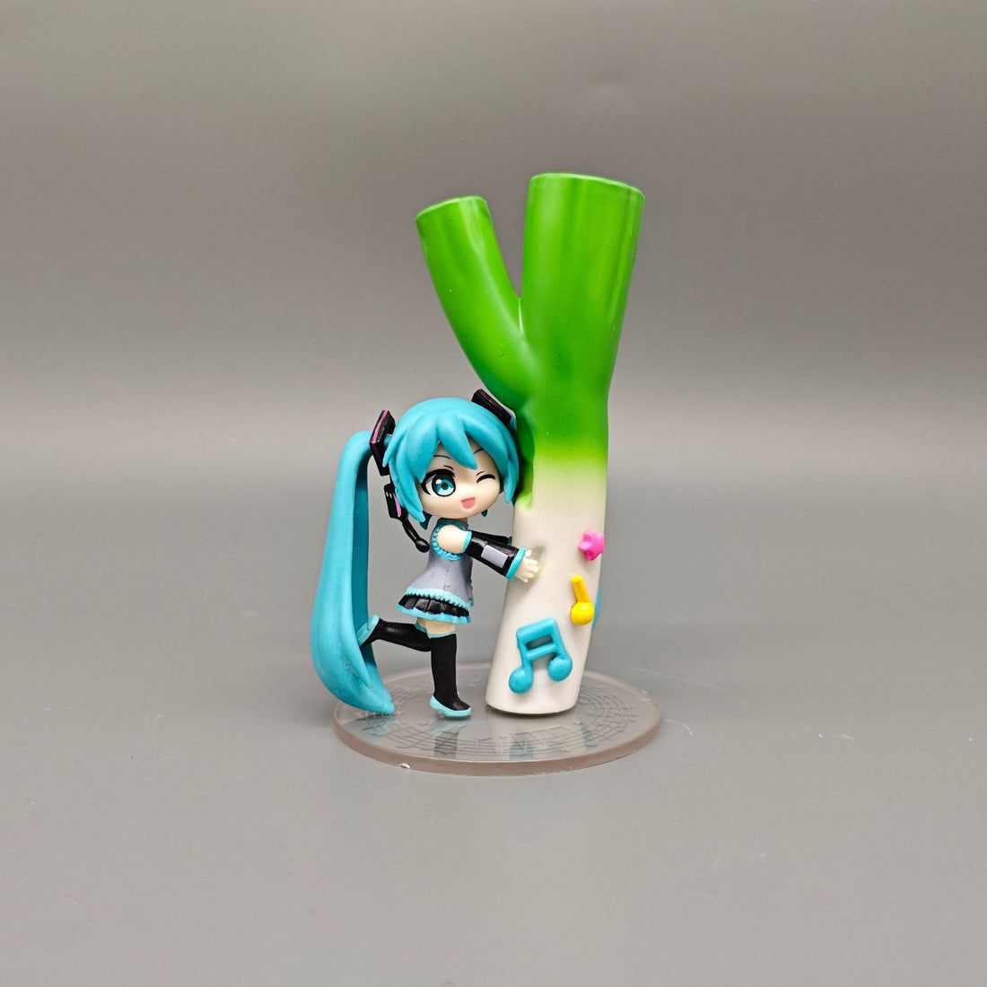 The Chibi Hatsune Miku Holding Green Onion 12cm PVC Figure playfully depicts the blue-haired character with headphones beside a large leek. Featuring a music note and star base, it embodies Hatsune Miku&