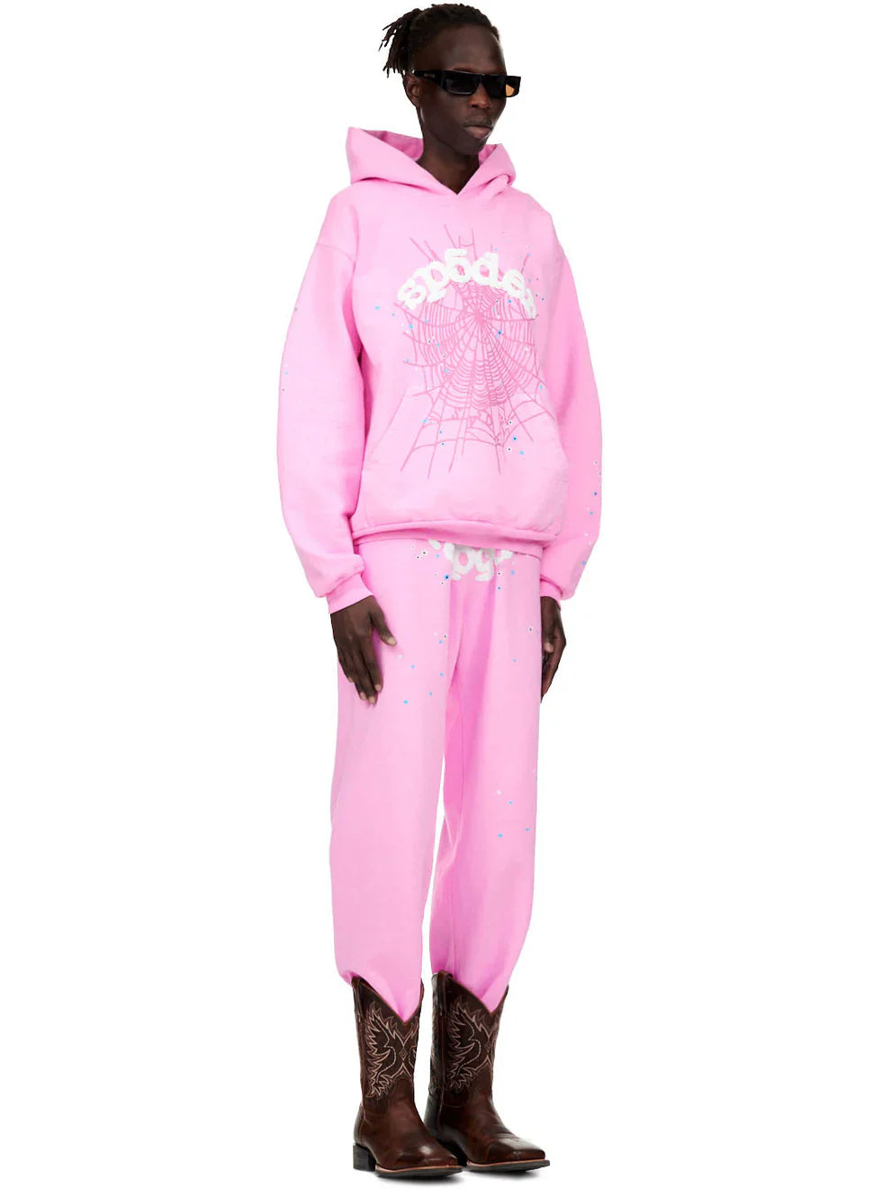 Wearing the Pink Graphic Hoodie with Spider Web Print – Comfort Fit Pullover Hoodie, pink pants with white designs, brown cowboy boots, and black sunglasses, a person stands against a plain white background.