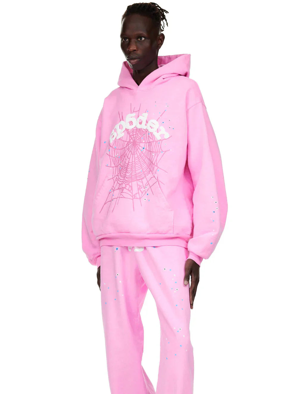 A person wears the Pink Graphic Hoodie with Spider Web Print, a comfort fit pullover adorned with bold white web graphics and stylized text, paired with matching pants featuring small white and blue speckles for a vibrant streetwear look.