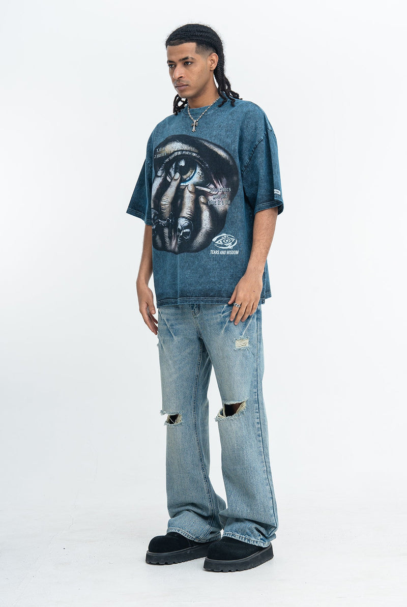Model wearing an oversized blue graphic tee with eye print, paired with distressed light jeans and black shoes
