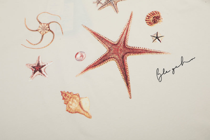 Coastal graphic T-shirt with large pink starfish, seashells, and beach-inspired botanical print on a cream background