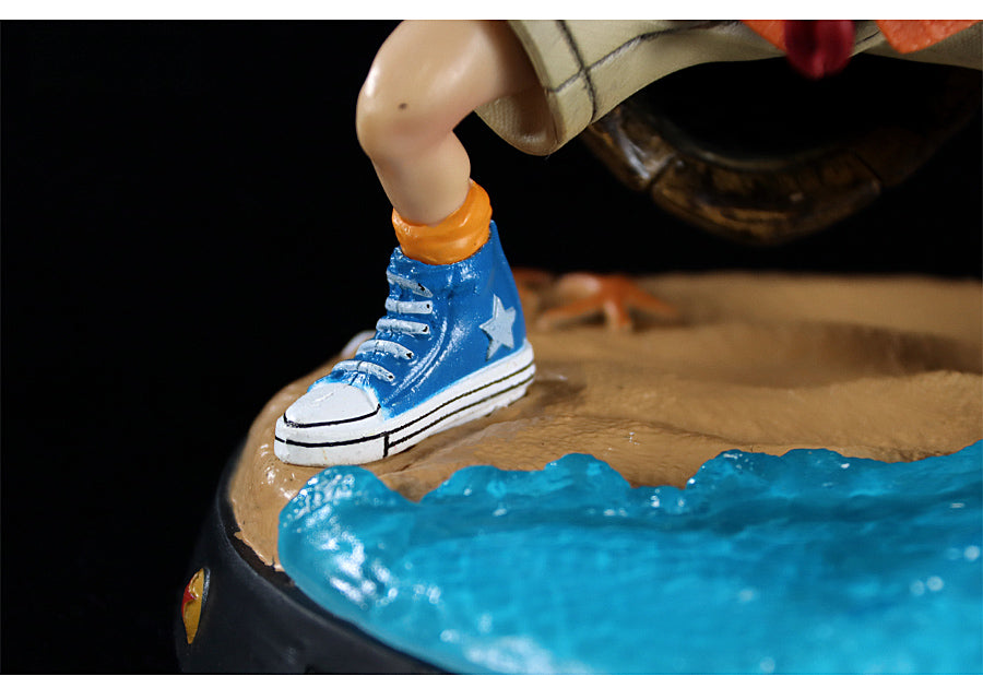 Close-up of the Seakoff Master Roshi Collectible Figure with Banana Fan captures its iconic style, featuring a blue sneaker with a white star, white laces, thick sole, and orange socks standing on sandy terrain near blue waves.